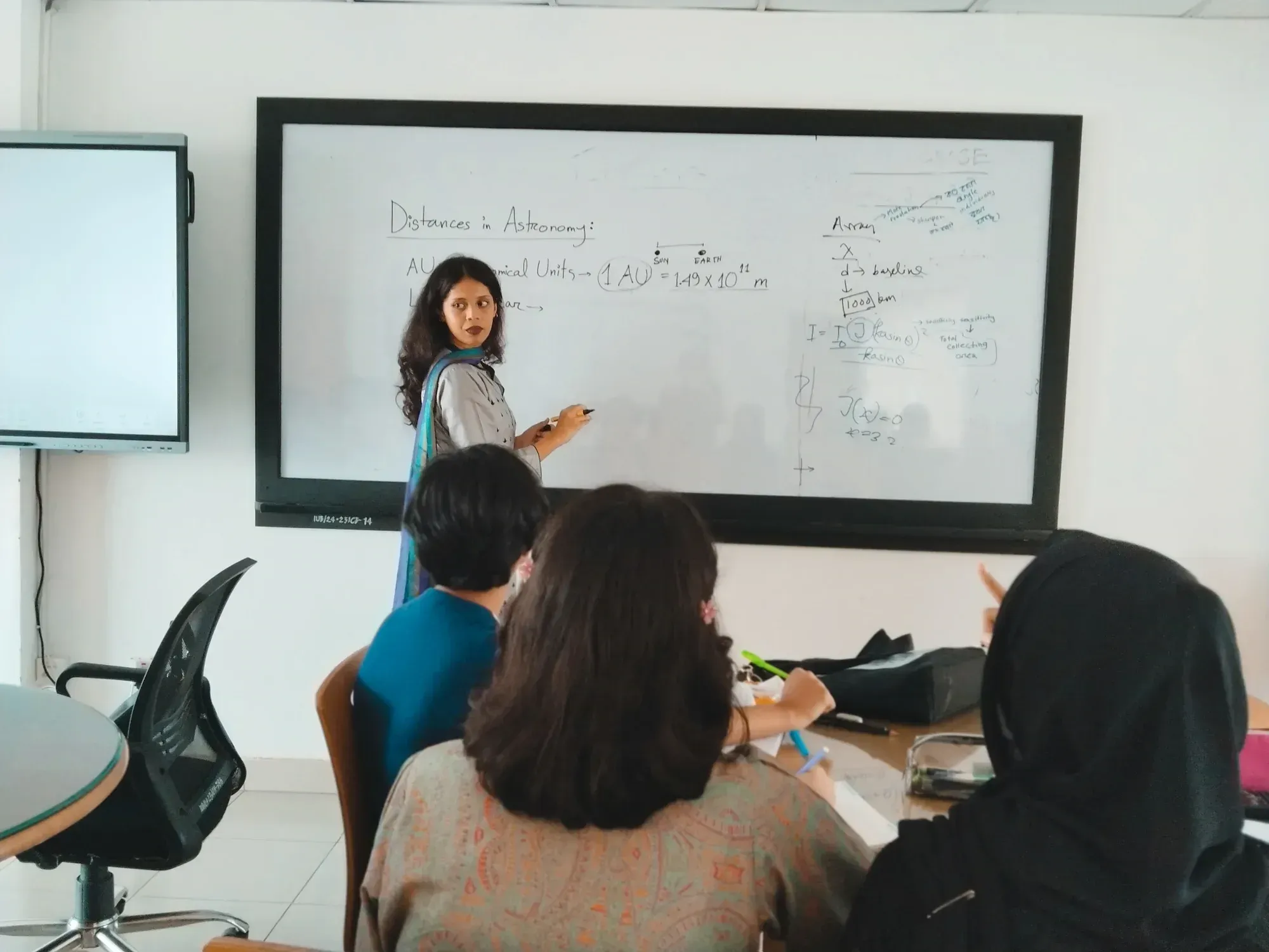 An instructor at the whiteboard beside the heading "Distances in Astronomy", with the astronomical unit written out as 1 AU = 1.49 × 10^11 m.