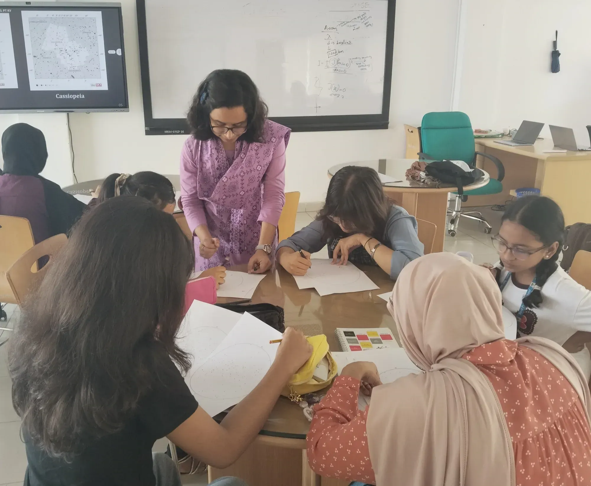 An instructor leans over a table guiding participants as they work on skymap sheets, with a chart of Cassiopeia displayed on the screen behind.