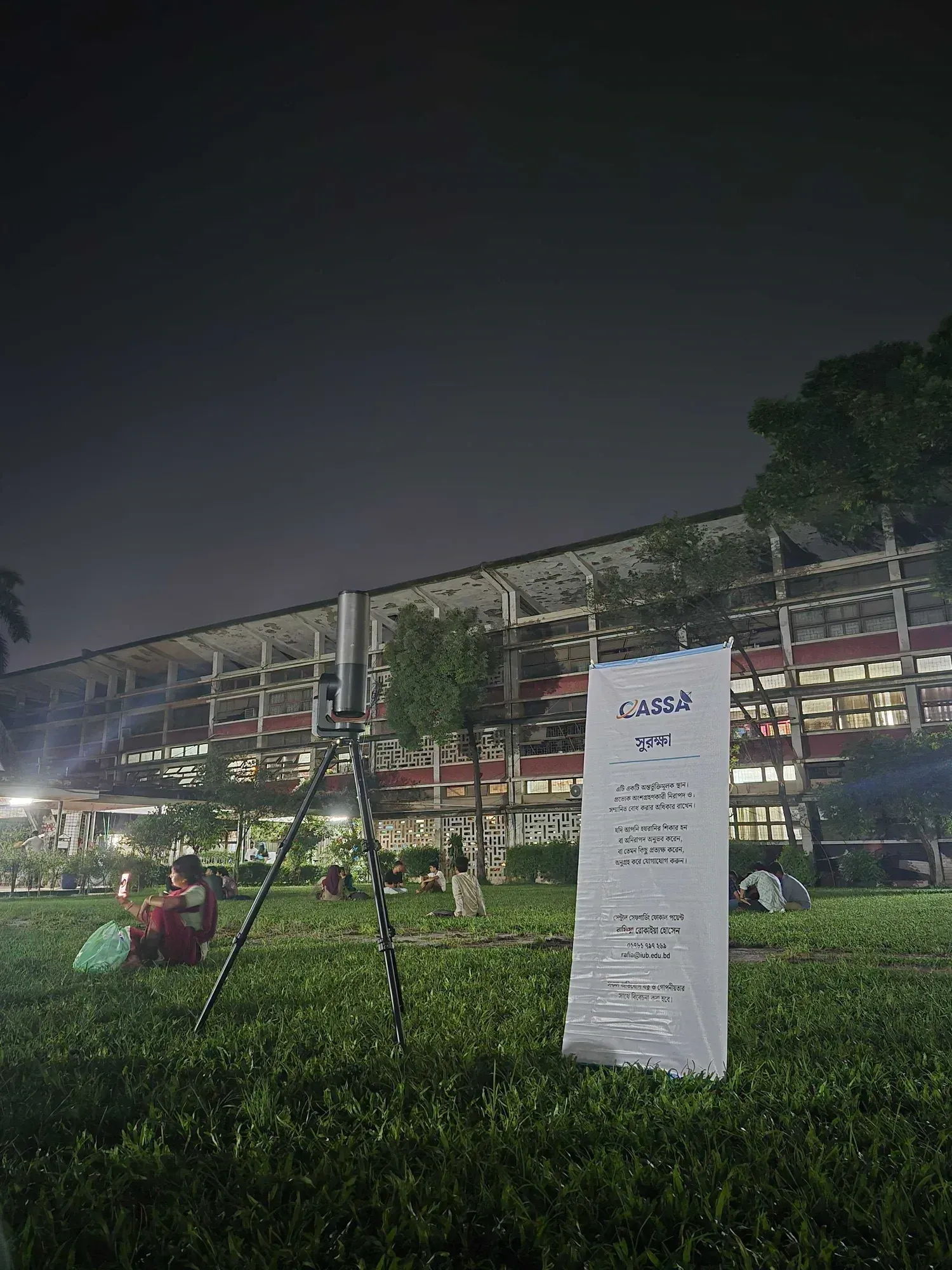 A telescope on a tripod beside a CASSA safeguarding banner on the TSC lawn, under a completely overcast night sky.