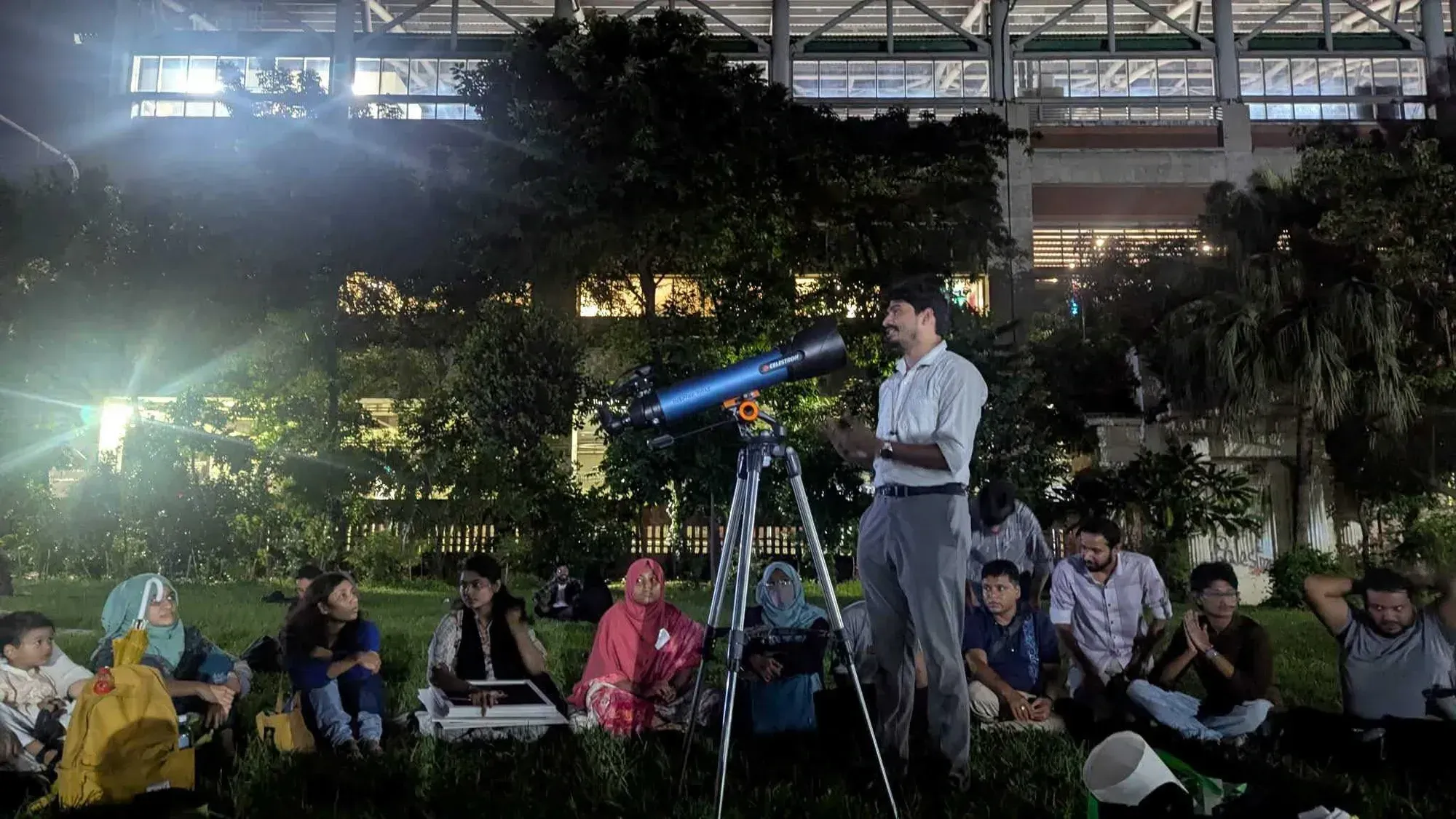 A participant standing beside a telescope, speaking to the seated audience on the TSC lawn.